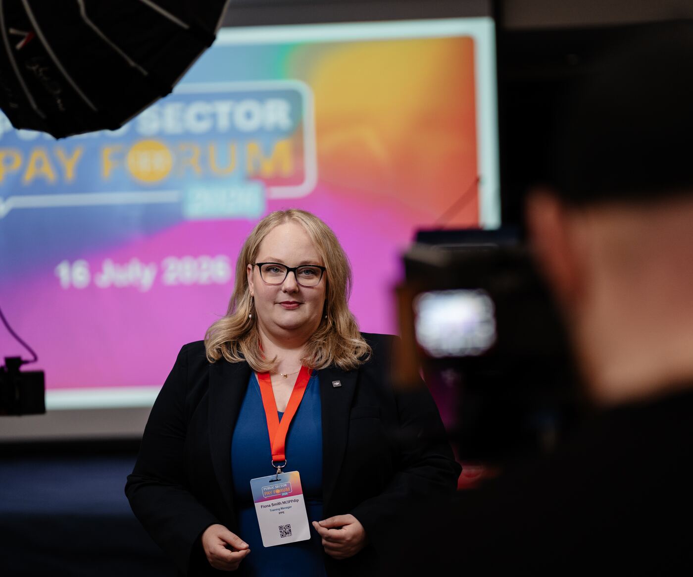 An interviewee in front of the Public Sector Pay Forum 2026 stage screen dated 16 July 2026, with the camera in the foreground